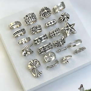 Op de afbeelding: Een collectie van tweeëntwintig zilverkleurige ringen en sieraden met diverse reliëf kruismotieven, bloemmotieven en gegraveerde tekst. Het assortiment bevat brede banden, open ringen en een groot kruisvormig hangerstuk.