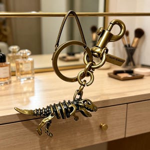 Peut inclure: Un porte-clés en métal doré vieilli avec une breloque en forme de squelette de dinosaure suspendue à un anneau circulaire et un mousqueton. La breloque représente la structure squelettique détaillée d'un dinosaure théropode.
