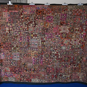 Puede incluir: Un gran tapiz rectangular que presenta un mosaico de cuadrados de tela multicolores. El tejido muestra bordados intrincados, patrones geométricos y pequeños espejos circulares reflectantes cosidos en toda la superficie de la pieza.