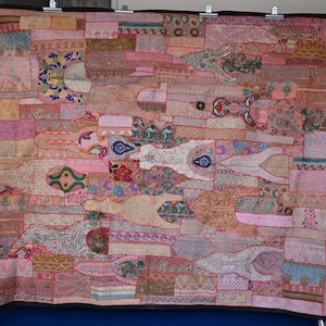 May include: A large rectangular wall hanging featuring a patchwork of pink, peach, and cream fabric segments. The textile is decorated with intricate embroidery, metallic thread accents, sequins, and diverse floral and geometric patterns throughout.