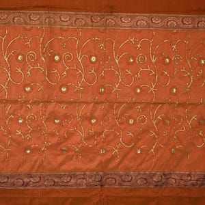 May include: A rectangular burnt orange textile featuring a central panel decorated with gold-colored embroidered swirling vine patterns and small circular sequins. The piece is framed by a darker brown border with a repeating floral motif.