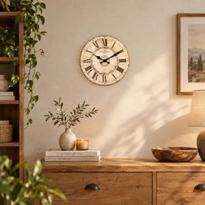 May include: A round wall clock with a distressed cream-coloured face, black Roman numerals, and black hands. The clock features a central circular design with text and markings, mounted on a plain wall above a wooden dresser with decor items.