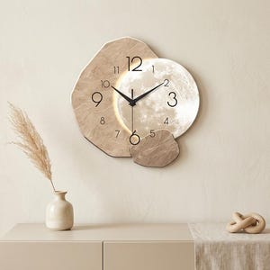 Peut inclure: Une horloge murale avec un cadre irrégulier superposé beige et crème, présentant un motif central circulaire rappelant la lune. Le cadran affiche des chiffres arabes noirs et des aiguilles fines, avec une lumière douce éclairant le motif.