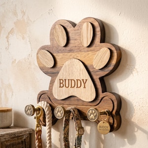 Könnte beinhalten: Ein wandmontierter Leinenhalter aus Holz in Pfotenform mit einer Basis aus dunklem Holz und helleren Holzakzenten. Das zentrale Ballenpolster ist mit dem Namen BUDDY graviert. Er verfügt über drei messingfarbene Haken mit Pfotenmotiven.