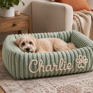 May include: A rectangular sage green pet bed with a ribbed, plush texture. The front side features the name Charlie embroidered in a cream-colored rope script, accompanied by a matching embroidered paw print design on the right side.