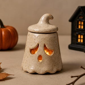 May include: A beige ceramic ghost-shaped jar with a speckled finish and a removable lid. The ghost features cut-out eyes and a mouth that emit a warm orange glow from within, creating a hollow vessel for storage or decorative lighting purposes.