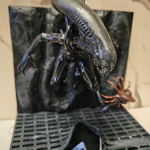 Op de afbeelding: Een zwart, glanzend Xenomorph-figuur dat uit een getextureerd wandpaneel tevoorschijn komt, met een langwerpig hoofd en scherpe ledematen. Een klein, bruin, veelpotig facehugger-wezen bevindt zich bij de basis op een zwart, gerasterd vloerplatform.