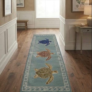 May include: A rectangular runner rug in a light blue hue featuring three sea turtles in blue, brown, and olive green. The rug is bordered by a white geometric wave pattern and finished with white fringe tassels on both short ends.