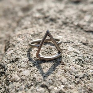 May include: A silver-coloured ring featuring a prominent open triangle design. The metal has a slightly oxidised, matte finish. The text 925SilvershopGifts is visible on the right side of the band. The ring is positioned upright on a textured surface.