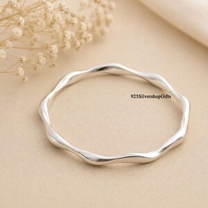 May include: A polished silver bangle bracelet with a continuous wavy, undulating design. The metal surface is smooth and reflective. The text 925SilvershopGifts is printed in black near the right side of the circular silver band.