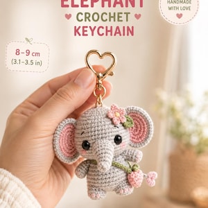May include: A gray crochet elephant keychain featuring pink inner ears, black eyes, and a small pink flower on its head. The elephant carries a tiny pink strawberry-shaped bag on a green strap. It is attached to a gold-colored heart-shaped clasp.