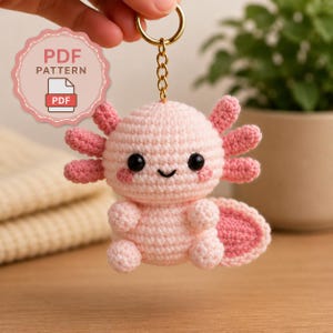 May include: A light pink crocheted axolotl keychain with dark eyes, a small smile, and three sets of darker pink external gills on each side of its head. A gold-toned metal chain and ring are attached to the top. A badge reads PDF PATTERN.
