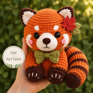 May include: A crocheted red panda plush toy featuring orange and dark brown yarn, a white muzzle, black eyes, a green bow tie, and a red maple leaf accessory on its head. A circular white label with the text PDF PATTERN is visible on the left.
