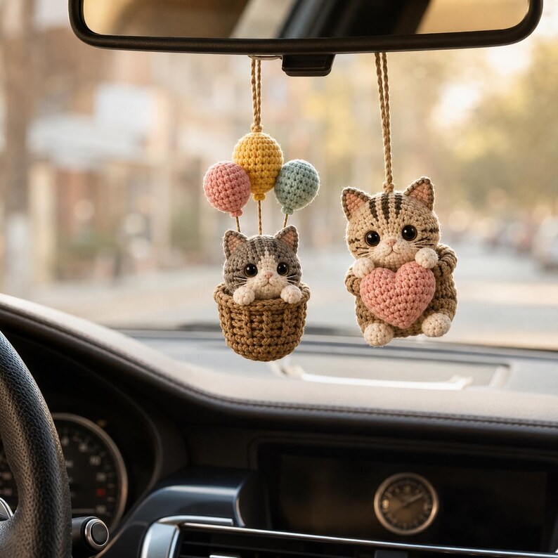 May include: Two crocheted cat ornaments hanging from a rearview mirror. One cat sits in a brown basket attached to three small balloons in pink, yellow, and blue. The other cat is brown with dark stripes and holds a pink crocheted heart in its paws.