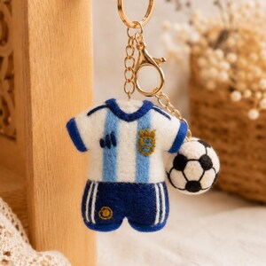 May include: A felt keychain featuring a miniature soccer uniform with a white and light blue striped jersey and dark blue shorts, paired with a small black and white soccer ball charm, all attached to a gold-toned metal ring and lobster clasp.
