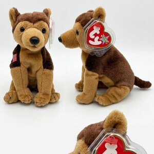 May include: A plush dog toy with tan and dark brown fur, featuring a black nose and eyes. The toy has a small American flag patch on its chest and a red heart-shaped Ty Beanie Babies tag attached to its ear, encased in a clear plastic protector.