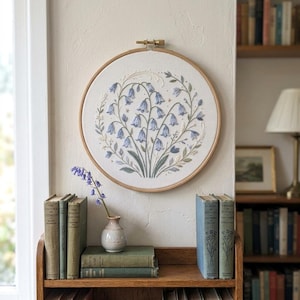 Könnte beinhalten: Ein runder hölzerner Stickrahmen mit weißem Stoff, der ein handgesticktes Motiv aus blauen glockenförmigen Blumen und grünen Blättern zeigt. Die Stickerei ist an einer Wand über einem Holzregal mit alten Büchern und einer kleinen Vase angebracht.
