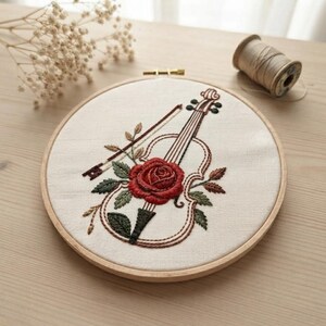 Puede incluir: Un bastidor de bordado que contiene una tela de color crema con el contorno cosido de un violín y un arco. Una rosa roja grande y detallada con hojas verdes está bordada sobre el centro del cuerpo del violín, acentuada por pequeñas enredaderas marrones.