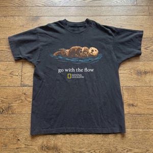 Może przedstawiać: Czarny t-shirt z krótkim rękawem z grafiką brązowej wydry morskiej unoszącej się w niebieskiej wodzie. Poniżej wydry znajduje się biały napis "go with the flow", a pod nim żółto-białe logo National Geographic umieszczone na środku klatki piersiowej.
