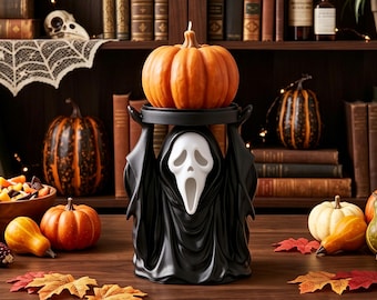 Puede incluir: Un calentador de cera negro con una figura blanca fantasmal de boca abierta que sostiene una bandeja circular sobre su cabeza. Una vela naranja con forma de calabaza descansa sobre la bandeja, creando un adorno decorativo de Halloween.