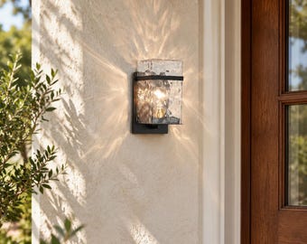 May include: A rectangular outdoor wall sconce featuring a black metal frame and a textured, clear glass shade. The light fixture is mounted on a light-colored stucco wall, emitting a warm, bright glow that casts distinct light patterns.