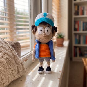 Pode incluir: Uma boneca de crochê com pele bronzeada, olhos grandes brancos e cabelo castanho. A boneca usa um boné de beisebol branco e azul com um emblema de pinheiro azul, uma camisa laranja, um colete azul, shorts cinza e tênis pretos e brancos.