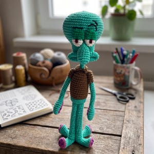 May include: A handmade crochet doll of a teal-skinned character with a large head, prominent nose, and brown shirt. The figure has white eyes with red pupils, dark eyebrows, and four teal legs with purple circular accents at the base of each foot.