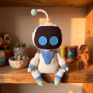 May include: A crocheted robot doll featuring a white body with light blue and mint green accents. The robot has a dark brown faceplate, two circular light blue eyes, and a single antenna with a mint green sphere on top, sitting in a seated pose.