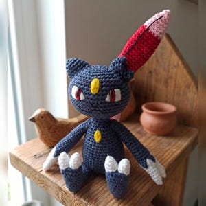 Op de afbeelding: Een donkerblauw gehaakt knuffeltje met een geel ovaal merkteken op het voorhoofd en de borst, puntige rode en roze oren, witte klauwen aan de handen en voeten, en rode ogen. Het figuurtje is gemaakt met duidelijk zichtbare garensteken.