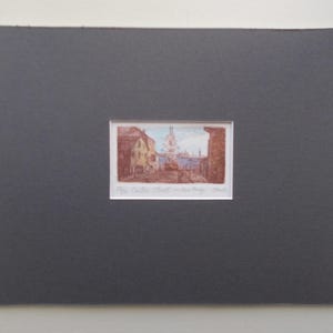 以下が含まれることがあります： 茶色、黄色、青色の建物が描かれた街並みの額入りプリント。 プリントは濃い灰色のマットボードにセットされています。 プリントの下部にはテキストがあります。