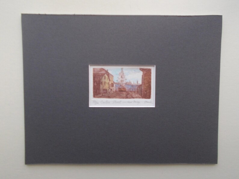 以下が含まれることがあります： 茶色、黄色、青色の建物が描かれた街並みの額入りプリント。 プリントは濃い灰色のマットボードにセットされています。 プリントの下部にはテキストがあります。