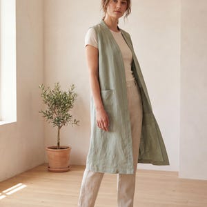 Peut inclure: Un long gilet sans manches vert sauge doté d'un devant ouvert sans col et de deux grandes poches plaquées sur le devant. Le vêtement est porté sur un t-shirt à manches courtes couleur crème et un pantalon beige clair à coupe droite.