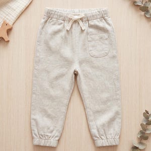 May include: Beige pants featuring an elastic waistband with a functional drawstring tie, a single patch pocket on the upper right side, and elasticized cuffs at the ankles. The garment is crafted from a textured, light-colored fabric.