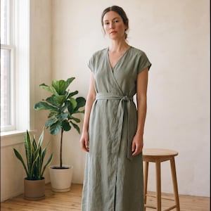May include: A sage green midi-length wrap dress featuring short sleeves, a V-neckline, and a matching fabric tie belt at the waist. The garment is constructed with a simple, clean silhouette and falls to a straight hemline below the knees.