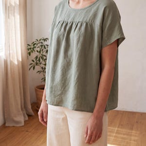 May include: A sage green short-sleeved blouse featuring a round neckline and a gathered yoke detail across the chest. The top has a loose, relaxed fit with a straight hemline and is paired with plain off-white trousers in a neutral colour palette.