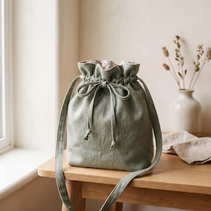 May include: A sage green drawstring bucket bag with a long shoulder strap. The exterior is a solid muted green fabric, whilst the interior features a floral pattern in shades of pink, red, and green. The bag is cinched at the top with a matching tie.