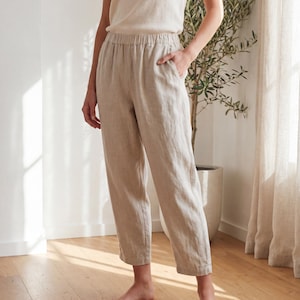 May include: Beige trousers featuring an elasticized waistband and a relaxed, straight-leg fit. These pants are crafted from a textured fabric and include side pockets, falling to a cropped length above the ankles with visible vertical seam detailing.