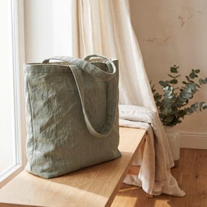 Peut inclure: Un sac fourre-tout vert sauge doté de deux anses et d'une doublure à motif floral visible. Le tissu extérieur présente un aspect texturé et légèrement froissé. Le sac est de forme rectangulaire avec une base plate et une ouverture supérieure.