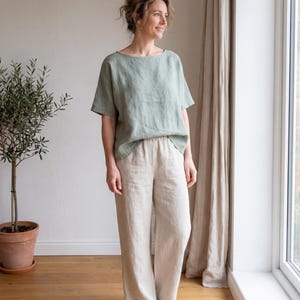May include: A sage green short-sleeved top with a wide neckline paired with loose-fitting beige trousers featuring an elastic waistband. The outfit consists of a relaxed-fit shirt and wide-leg pants, both crafted from a textured fabric, creating a simple ensemble.