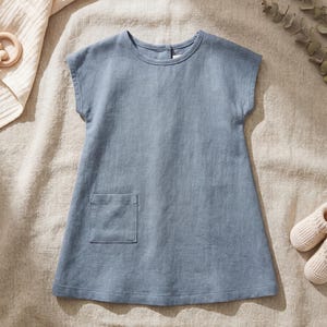 May include: A dusty blue sleeveless dress featuring a round neckline, a single patch pocket on the front, and a button closure at the back of the neck. The garment is constructed from a textured fabric with a simple, straight-cut silhouette.