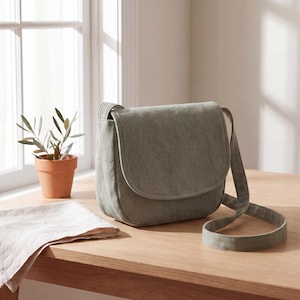 Puede incluir: Un bolso bandolera de color verde salvia con cierre de solapa redondeada y correa ajustable a juego. El bolso presenta una construcción de tela texturizada con líneas limpias y una silueta estructurada, diseñado para llevar objetos personales.