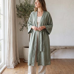May include: A sage green open-front robe featuring wide three-quarter length sleeves and a matching fabric tie belt at the waist. The garment is worn over a cream-colored tank top and wide-leg trousers, creating a layered look with a simple silhouette.