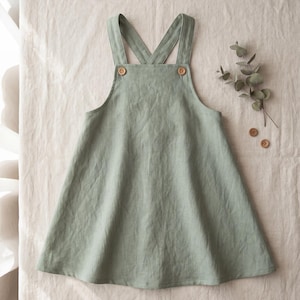 Peut inclure: Une robe chasuble vert sauge avec une jupe évasée, une encolure droite et de larges bretelles croisées dans le dos. Le vêtement est fixé par deux boutons en bois brun clair sur le devant, placés de chaque côté du corsage de la robe.