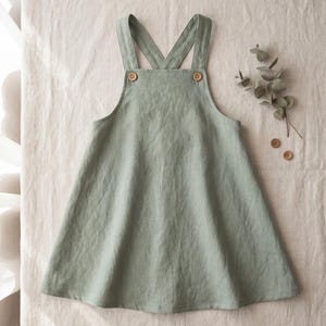 May include: A sage green pinafore dress featuring a flared skirt, a square neckline, and two wide shoulder straps that cross at the back. The garment is secured at the front with two small, round, light brown wooden buttons at the strap attachments.