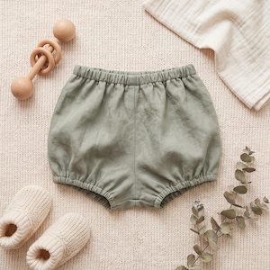 May include: Sage green baby bloomers with an elasticated waistband and leg openings, accompanied by a wooden rattle, a pair of cream-colored knit booties, a folded muslin cloth, and a sprig of dried eucalyptus leaves on a textured fabric surface.
