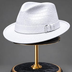 May include: A white fedora hat with a textured crocodile skin pattern, featuring a matching white hat band and a silver-toned metal buckle detail on the side, displayed on a gold and black marble stand.