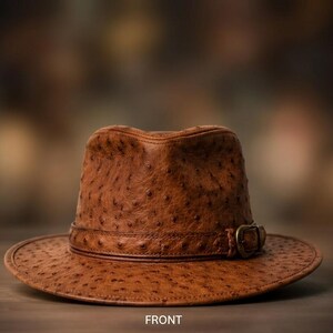 Pode incluir: Um chapéu fedora castanho com um padrão texturizado semelhante ao de avestruz na copa e na aba. O chapéu inclui uma faixa castanha a condizer à volta da base da copa, fixada com uma fivela metálica na lateral. A palavra FRONT está escrita abaixo.