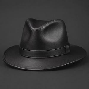 Puede incluir: Un sombrero fedora negro sólido que presenta una corona estructurada con un pliegue central y un frente pellizcado. El sombrero incluye una banda negra a juego que rodea la base de la corona, sobre un ala ancha y plana con un borde acabado.