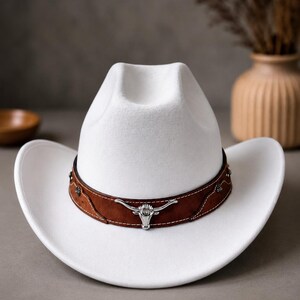 Puede incluir: Un sombrero de vaquero de fieltro blanco con una banda de cuero marrón ancha alrededor de la copa. La banda está decorada con costuras blancas, pequeños remaches plateados y un emblema central de metal plateado con forma de cráneo de longhorn.