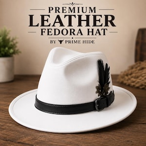 Pode incluir: Um chapéu fedora branco com uma faixa de couro preta com fivela e um detalhe decorativo de pena preta. O texto acima do chapéu diz PREMIUM LEATHER FEDORA HAT BY PRIME HIDE. O chapéu tem uma copa estruturada e uma aba larga e plana.